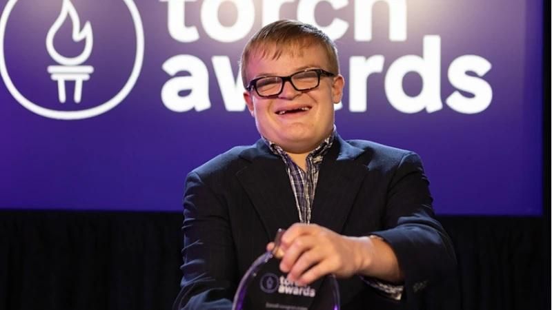 Travis Glock standing with the TORCH Awards logo in the background holding her award.
