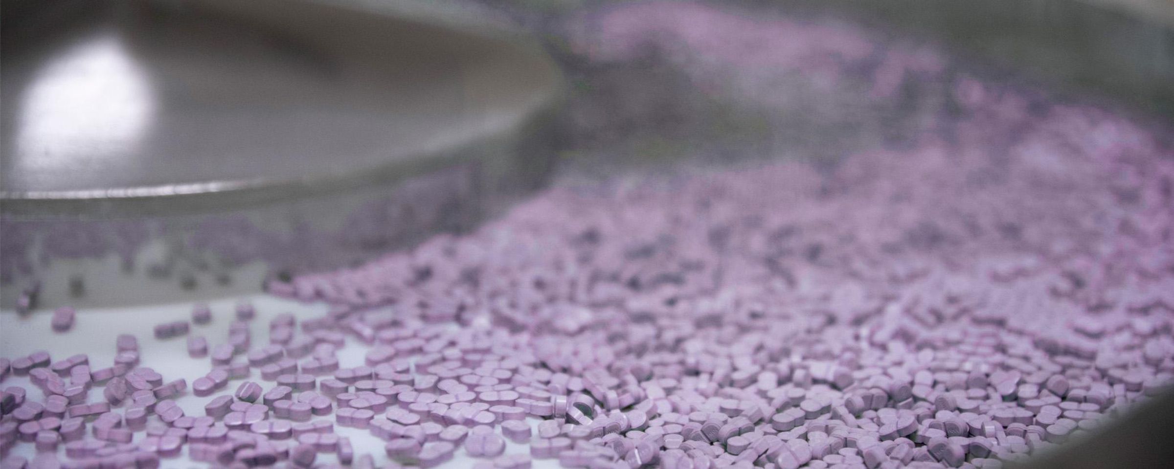 A stainless steel machine with a conveyor belt moves hundreds of purple pills along a Sanofi manufacturing line.