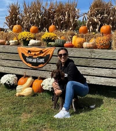 Melissa kneels ith her arms around her brown dog in a pumpkin patch.
