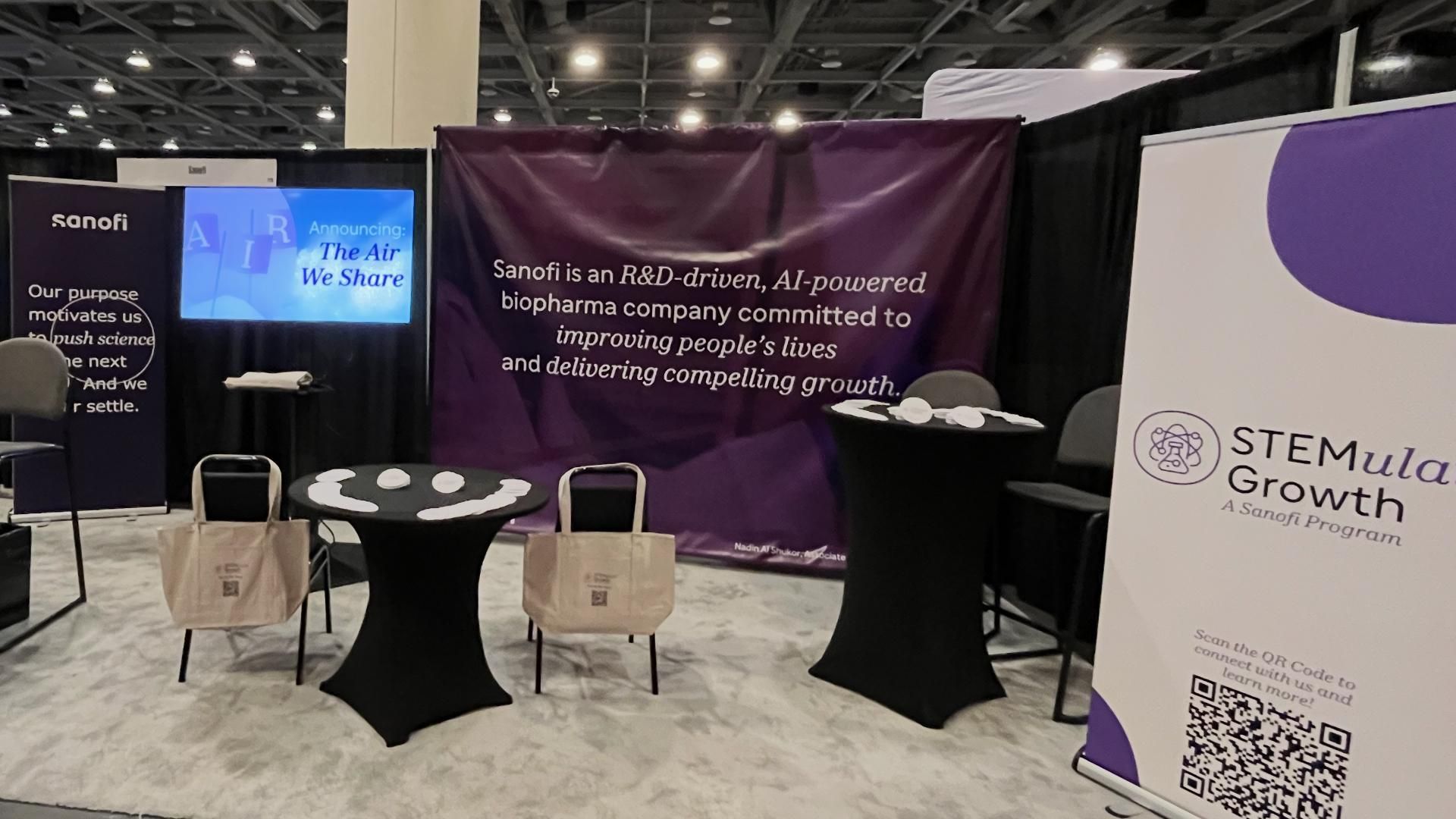 Sanofi conference booth with banners displaying “The Air We Share” and “STEMulate Growth” programs, tables with tote bags, and messaging about science-driven innovation and improving lives.