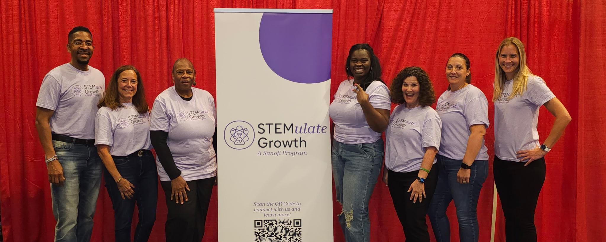 Seven people in matching purple “STEMulate Growth” t-shirts stand smiling beside a program banner with a red curtain backdrop.