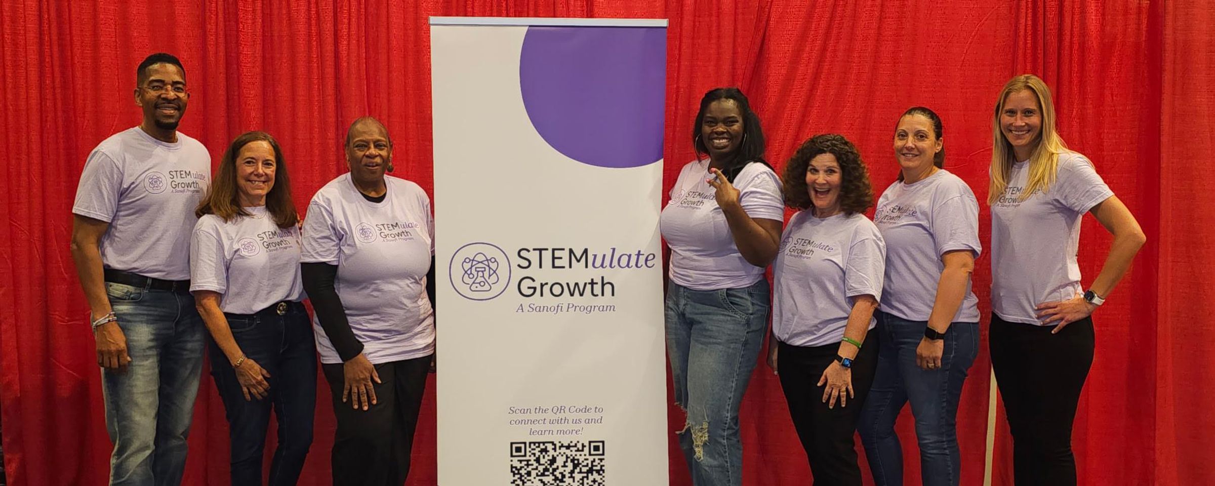 Seven people in matching purple “STEMulate Growth” t-shirts stand smiling beside a program banner with a red curtain backdrop.