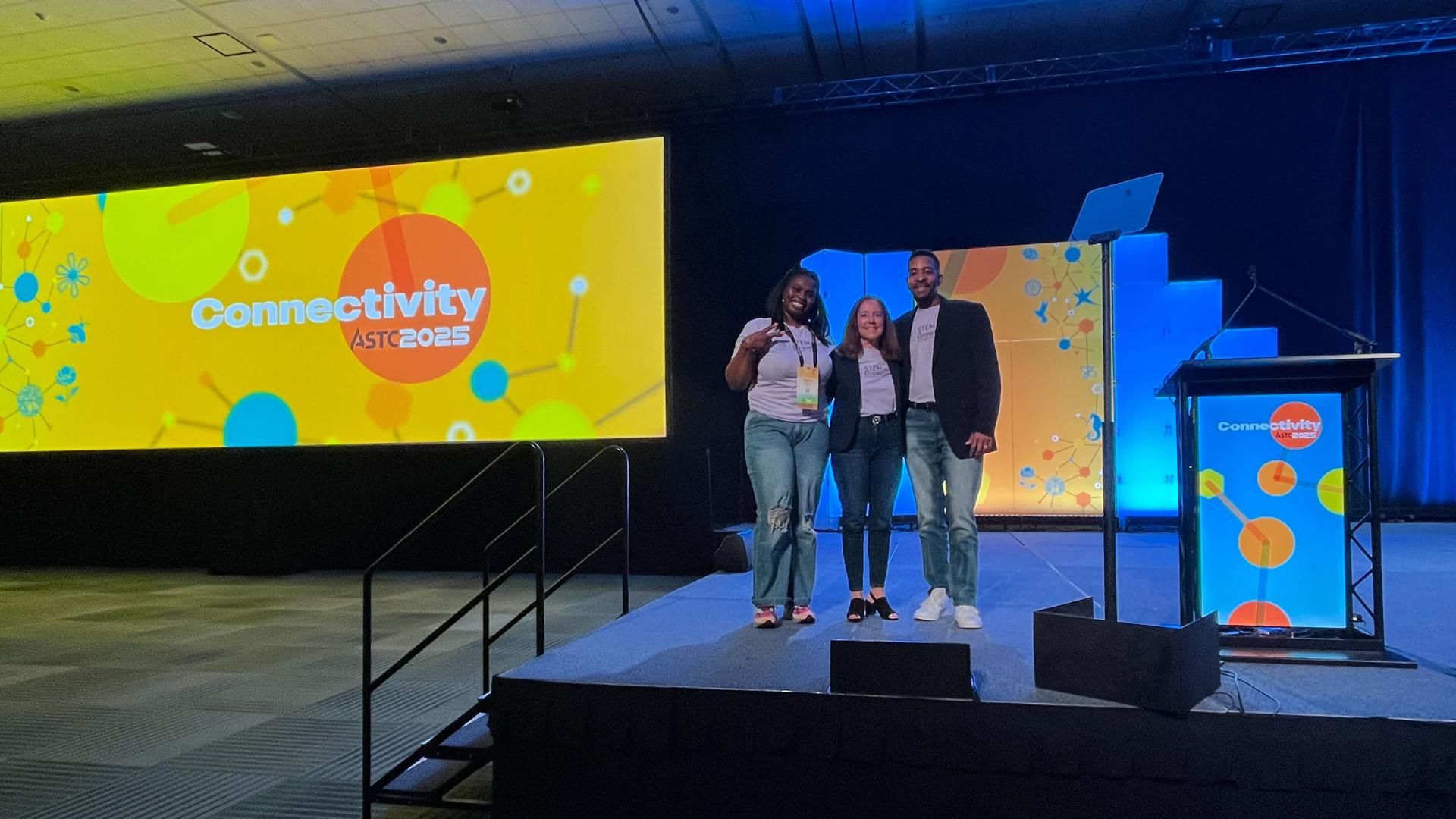 Three Sanofi representatives stand together smiling on stage at the ASTC 2025 Connectivity conference, with a large yellow conference screen and podium in the background.