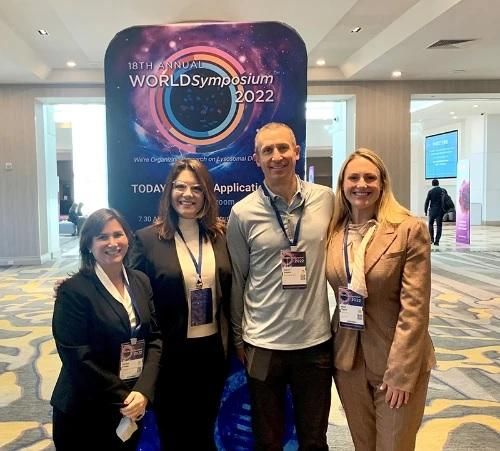 Kate and 3 attendees from various organizations smile at the camera with a banner for the 2022 Annual World Symposium behind them.