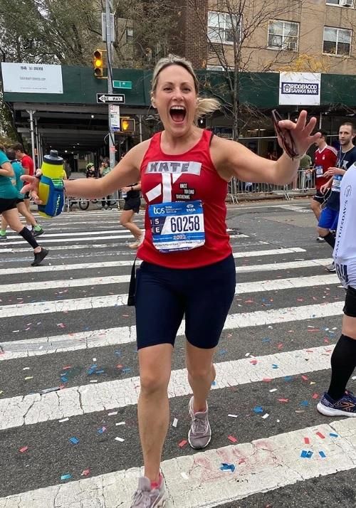 Kate in running gear smiles widely and throws out her arms at the camera midway through her third NY City Marathon.