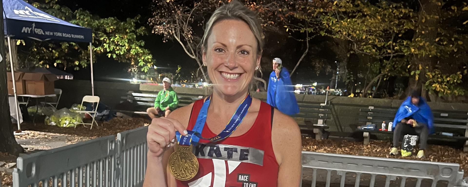 Kate Tighe wearing a red race shirt holds up her NY City marathon finshers medal.