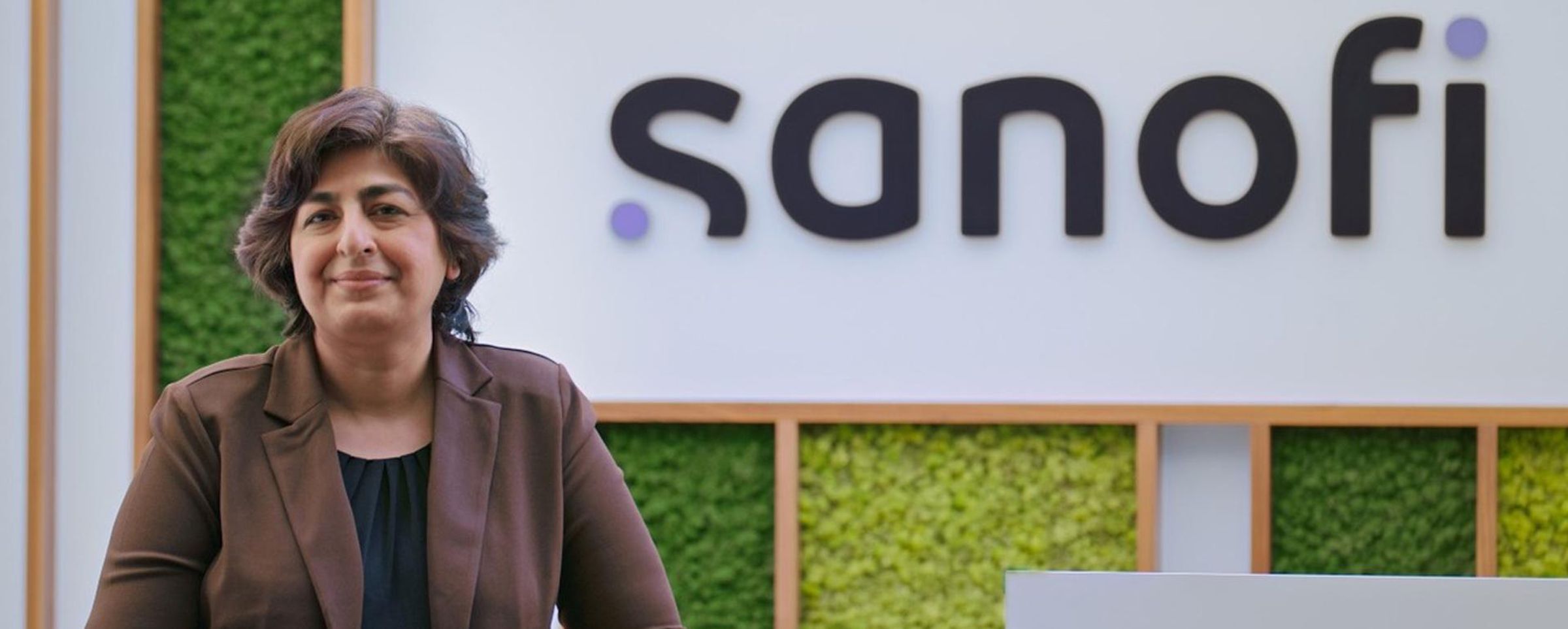 Ekta Seth Chhabra smiles at the camera while stadning in front of a living wall with a Sanofi logo.