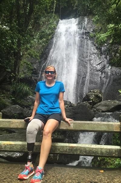 Melanie Stutts wears a bright blue shirt and sits on a railing in front of a waterfall in Hawaii.