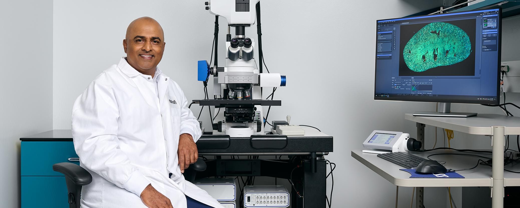 Scientist in white lab coat smiling while working at a microscope station with a computer monitor displaying green microscopic imagery.