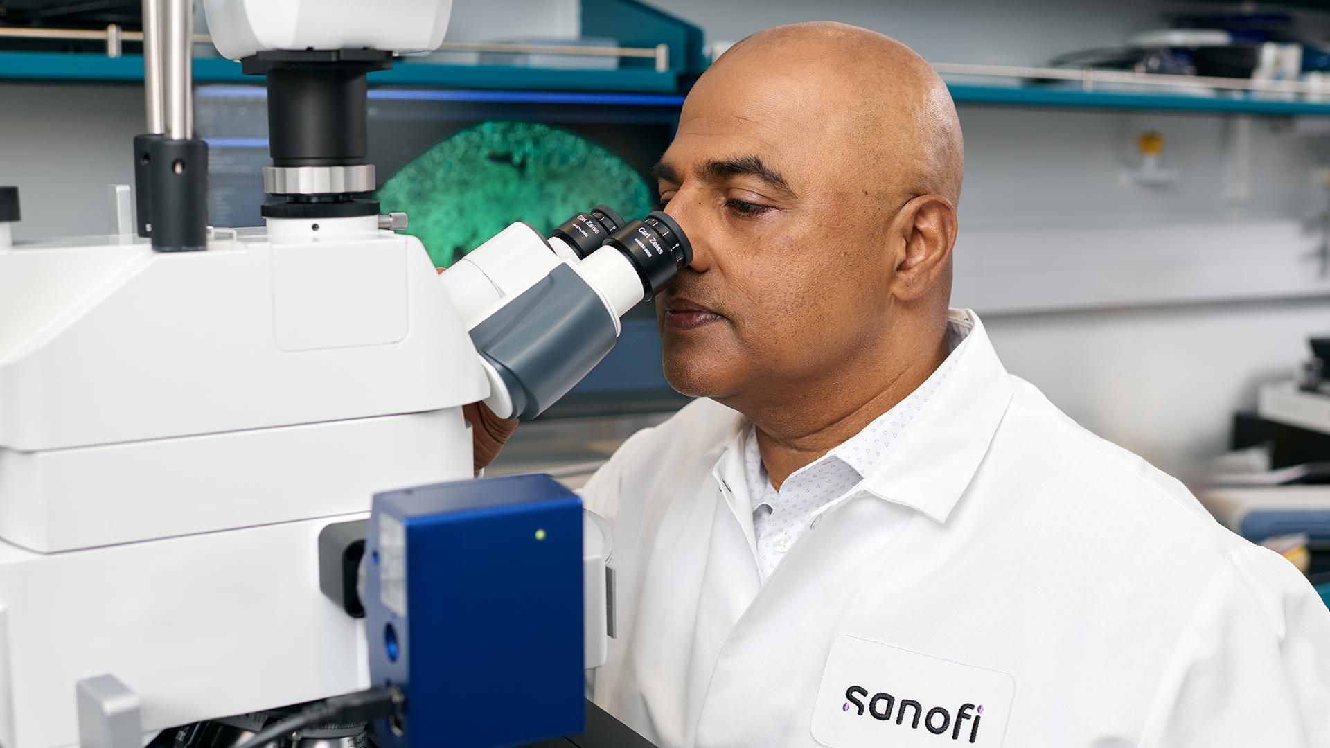 Srini Rao, wearing a white Sanofi lab coat, examines a sample using a microscope in a laboratory filled with scientific equipment.