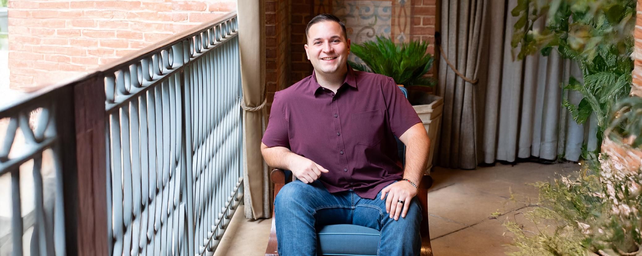 John, wearing a maroon shirt and jeans sits in a chair and smiles at the camera