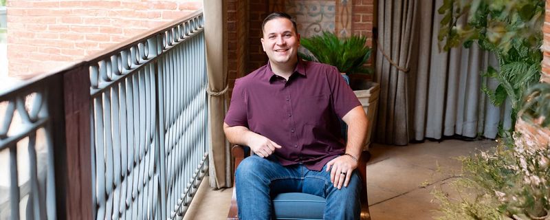 John, wearing a maroon shirt and jeans sits in a chair and smiles at the camera