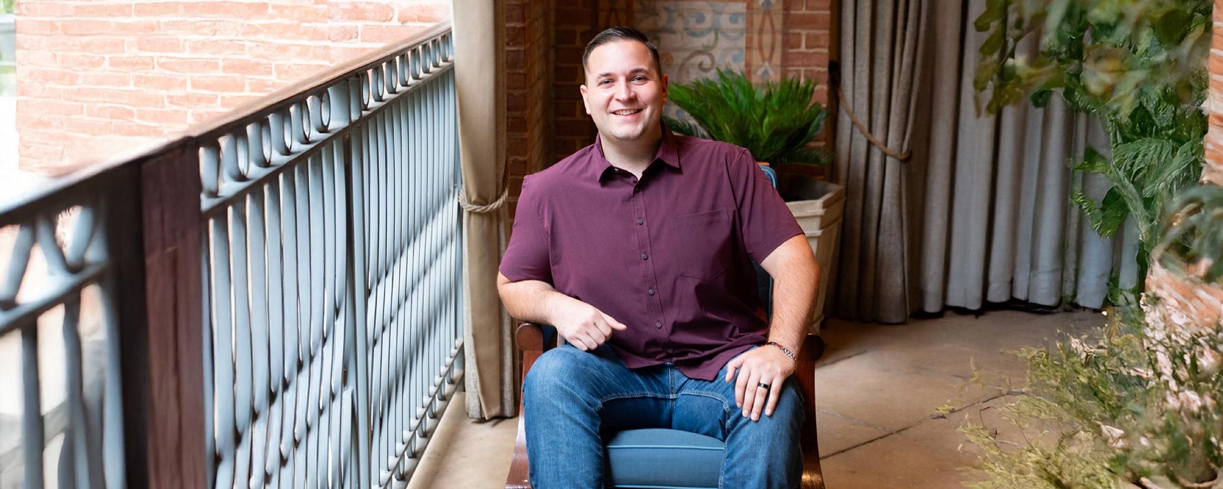 John, wearing a maroon shirt and jeans sits in a chair and smiles at the camera