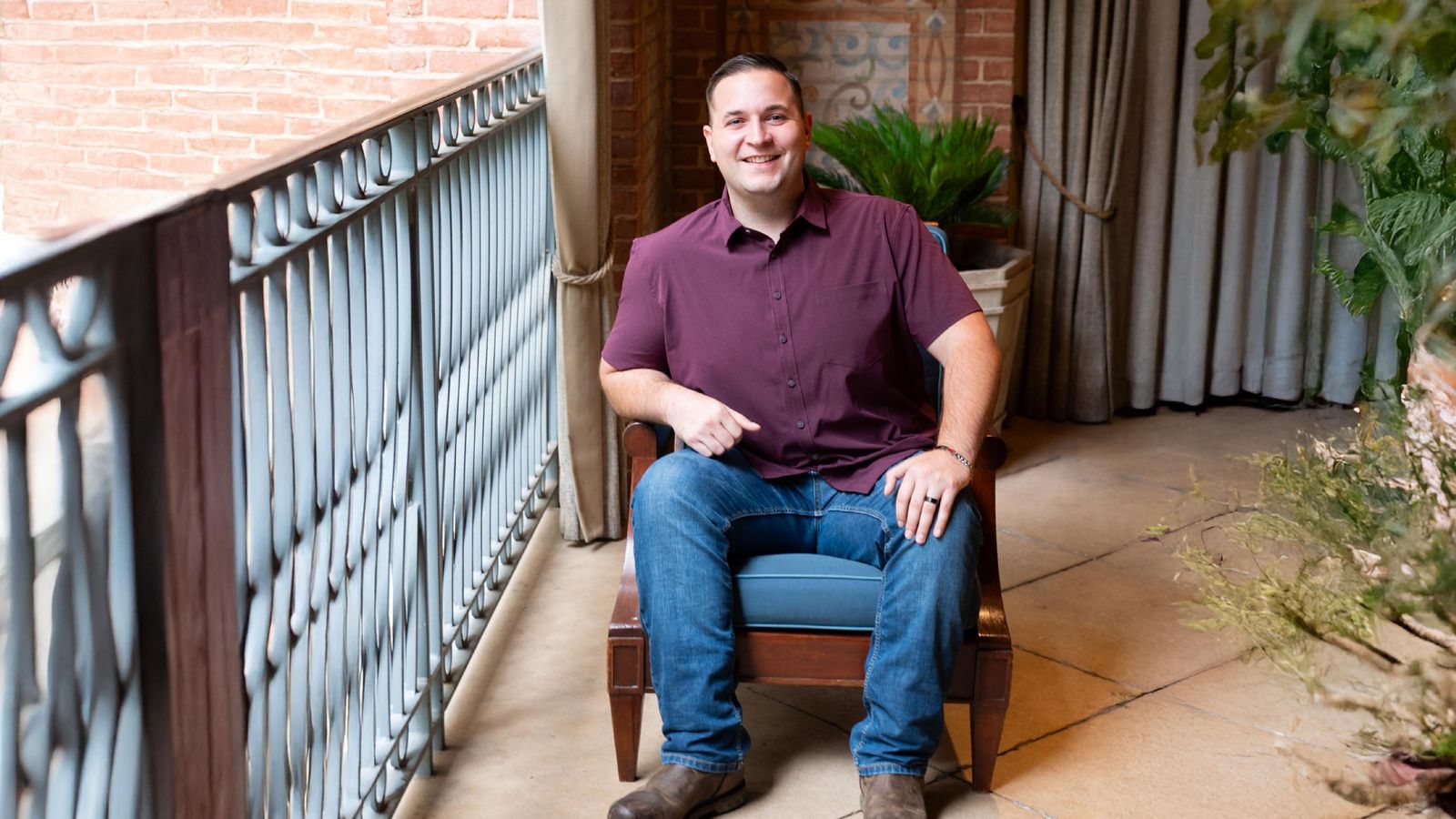 John, wearing a maroon shirt and jean sits in a chair and smiling at the camera