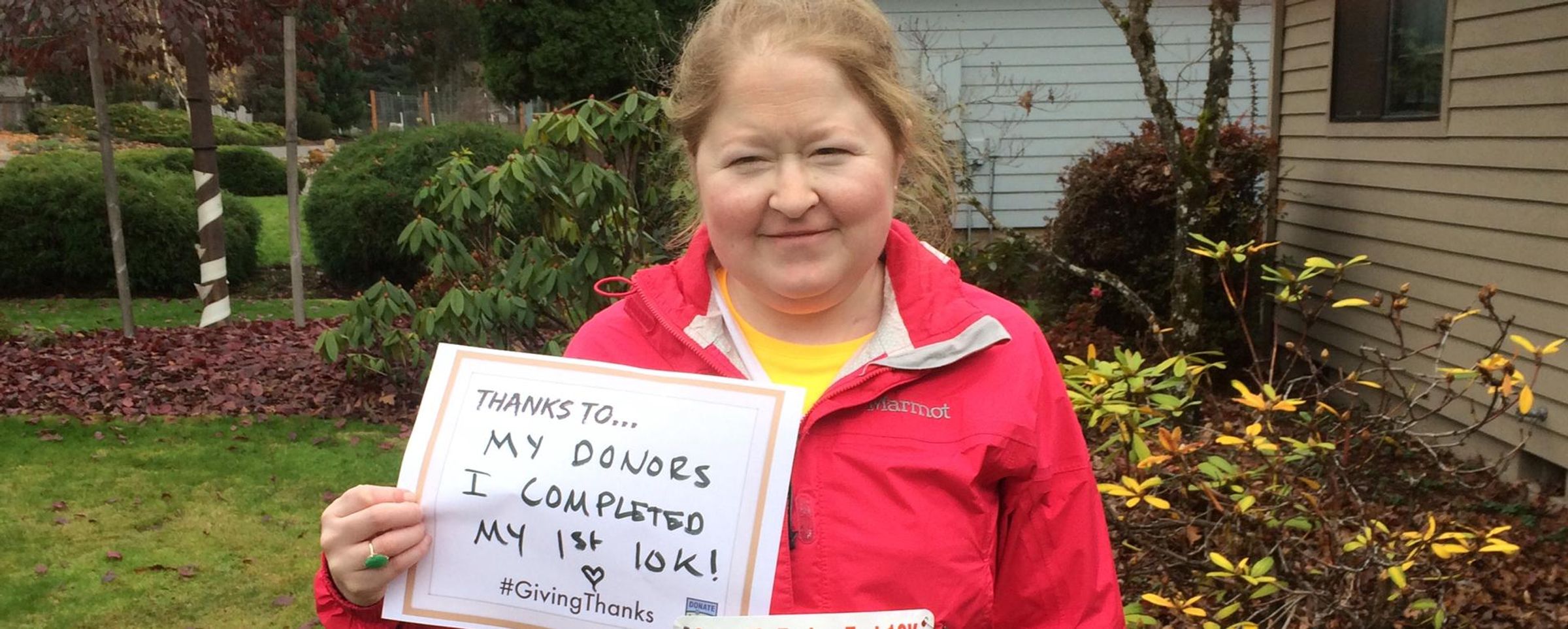 Aimee stands in a red coat holding a sign that says, "Thanks to my donors, I completed my first 10K!"