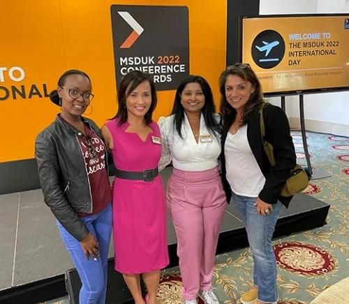 Rakhi Agarwal and three female colleagues stand in a link in front a step and repeat banner at a conference.