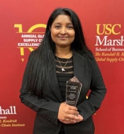 Rakhi Agarwal stands against a red step and repeat background holding a glass award for supplier diversity.
