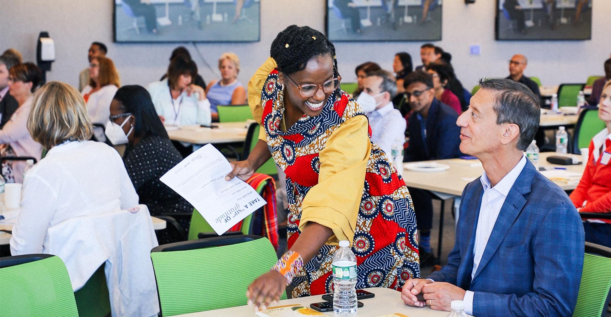 Nancy Ngotho in a patterned dress smiles at George Coutros and hands him a stack of papers.