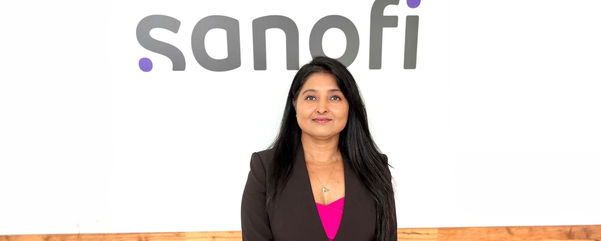 Rakhi Agarwal smiles at the camera while standing against a wall with the Sanofi logo.