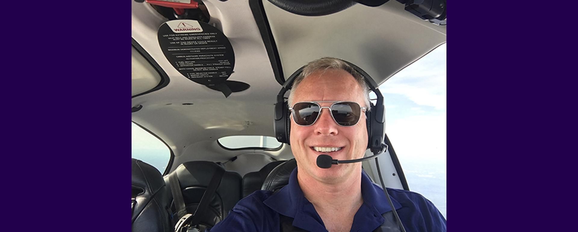 Will Richmond sits in the cockpit of his airplane mid-flight with sunglasses and a headset on, smiling at the camera.