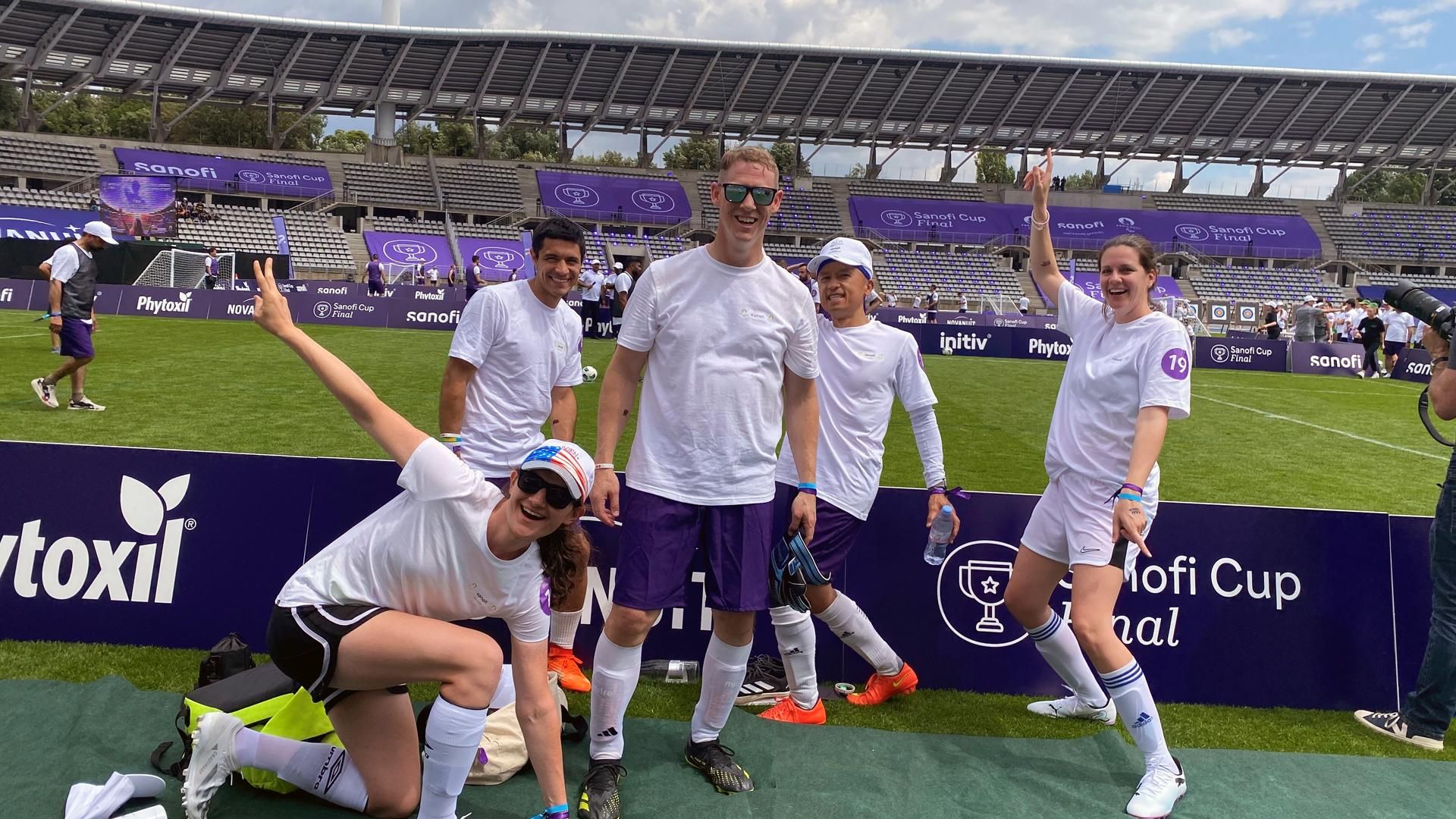 Josh Hellwig stands in the center of four of his teammates, who all smile and wave at the camera in their white Sanofi Cup t-shirts.