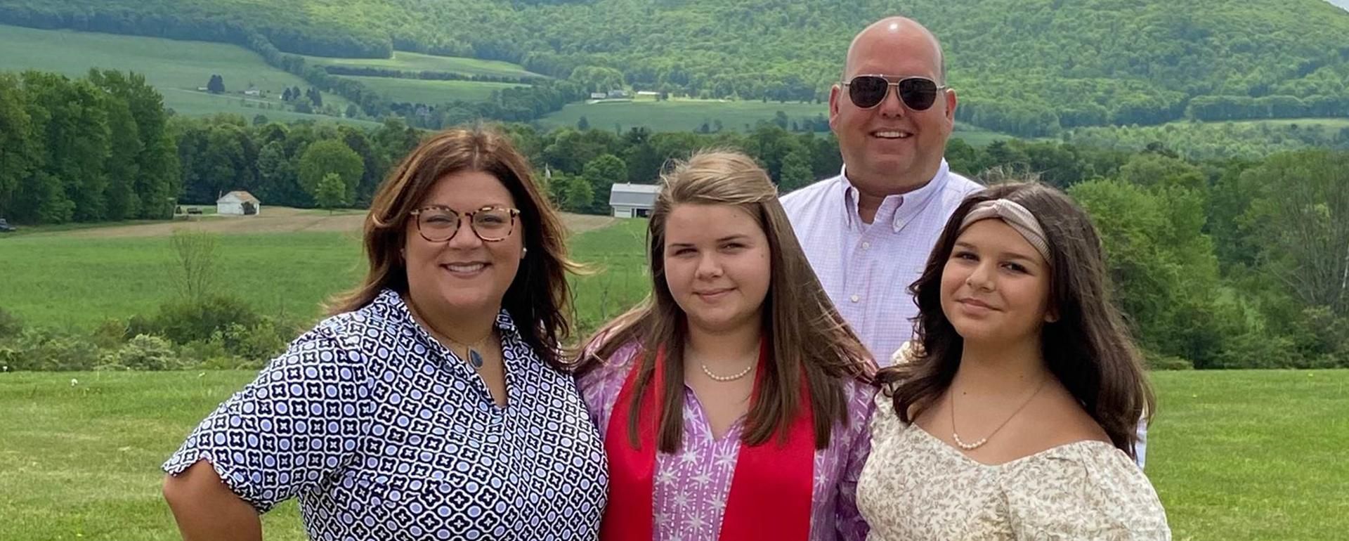 Lisa Moher stand with her husband and two daughters in a grassy field smiling at the camera.