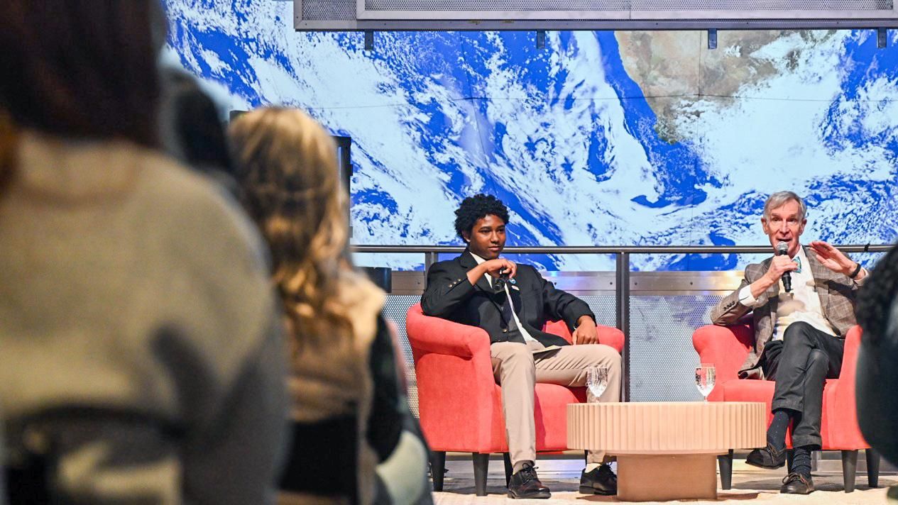 A Boys &amp; Girls Club student sits on a stage interviewing Bill Nye the Science Guy at the Museum of Science in Boston in front of a live audience, discussing air quality and youth engagement.