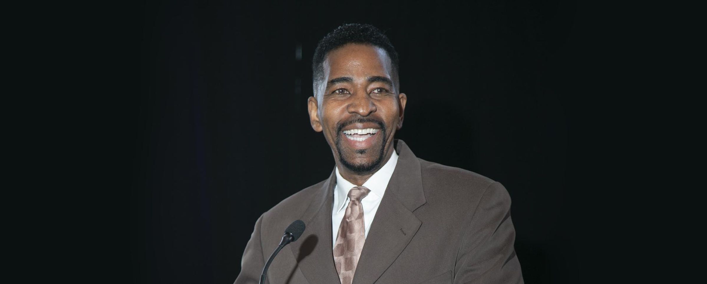 Phillip St. James, an african american man, stands against a black background, smiling at a crowd off screen as he speaks at a fundraiser.