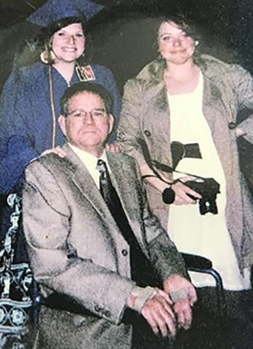 Amy Richards in a graduation cap and gown stnds with her father who is in a wheelchair, and her sister who holds a camera.