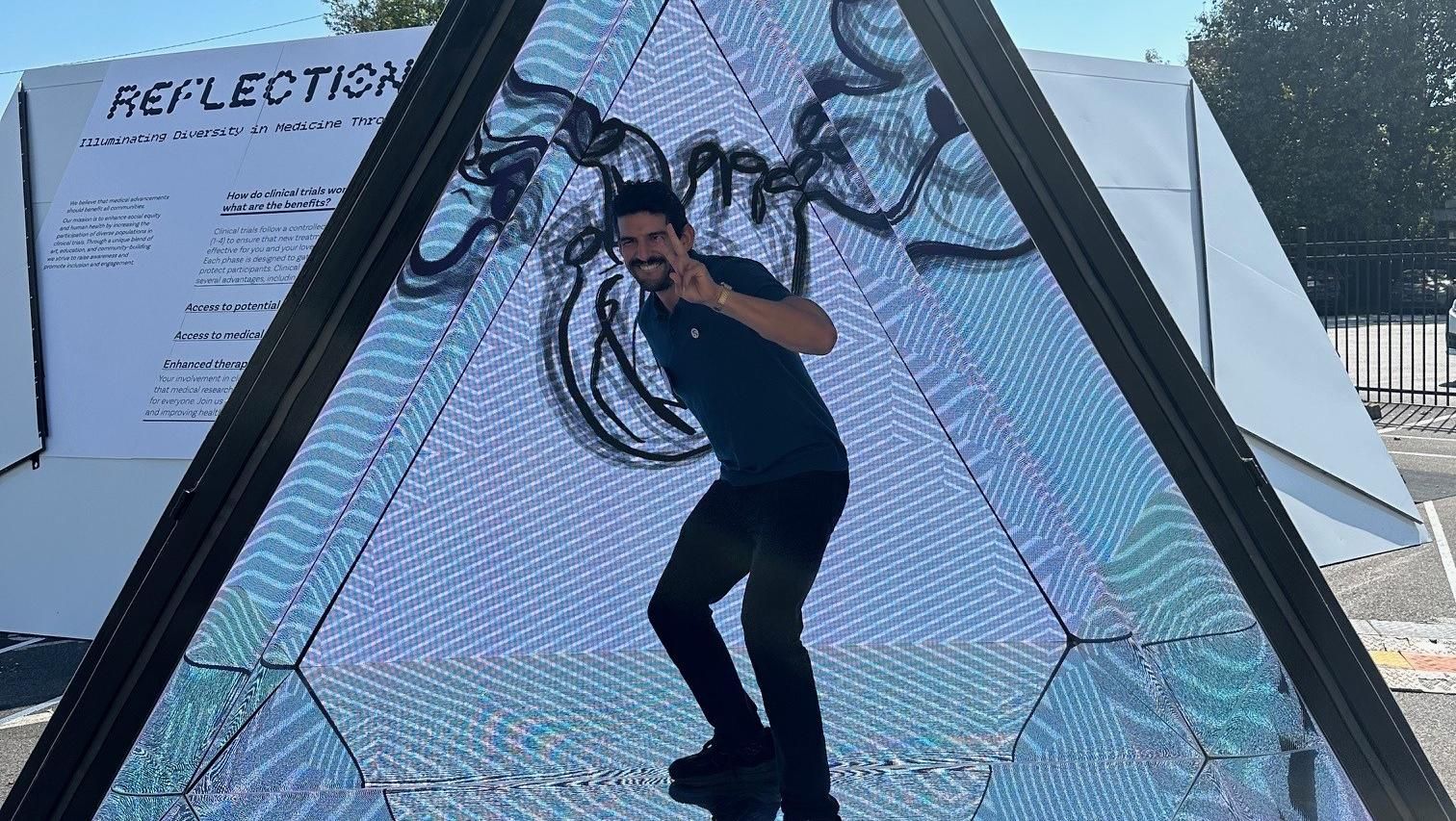 Albert Maffei, Community Outreach and Engagement Manager at Sanofi, dressed in a blue t-shirt, stands under a pyramid-shaped mirror art installation.