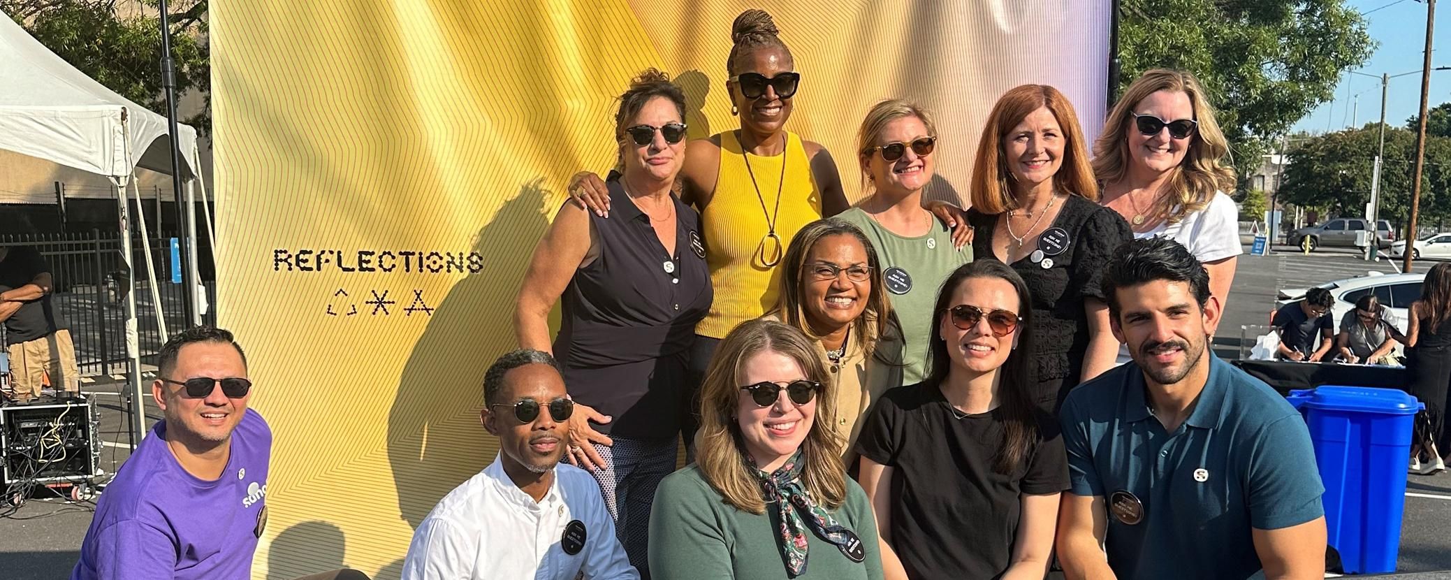 A group of the Sanofi team in t-shirts and jeans pose for a photo outside in front of a large tree.
