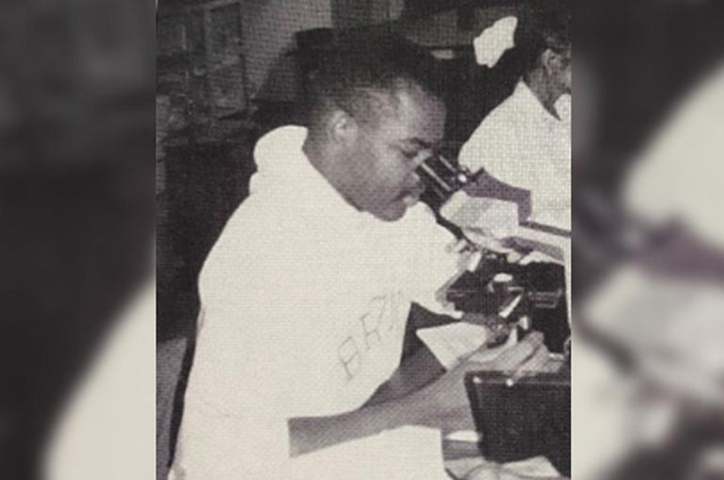 A black and white image of Corey Robertson looking into a microscope.