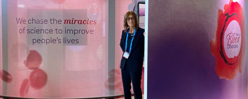 Dr. Karin Knobe, dressed in a suit with a lanyard, is standing in a medical conference booth that has images of red blood cells; on the left, there is text that says, “We chase the miracles of science to improve people’s live”; on the right, there is text that says, “Rare Blood Disorders.”