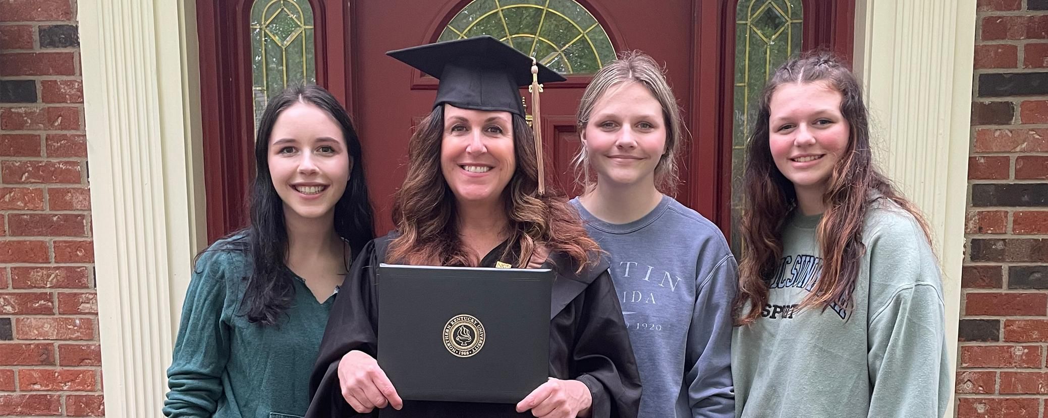 Katie stands in front of her house with her three daughters while wearing her graduation cap and gown holding her diploma.
