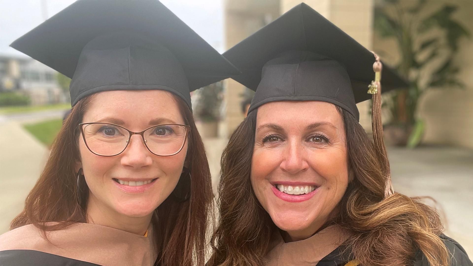 Katie and her friend smile at the camera while wearing their graduation caps and gowns after graduation.