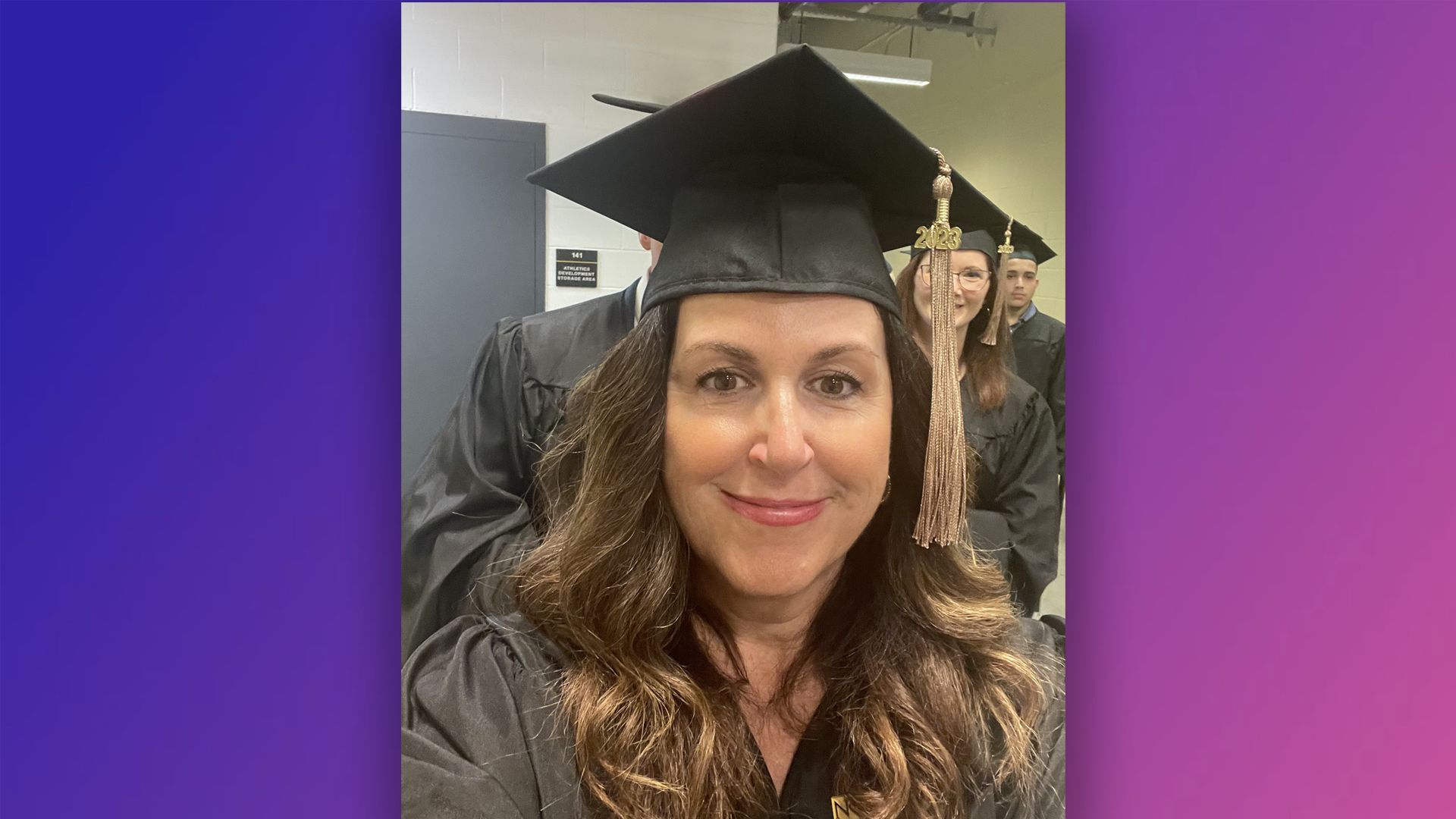 Katie smiles at the camera while wearing her graduation cap and gown waiting to walk for graduation.