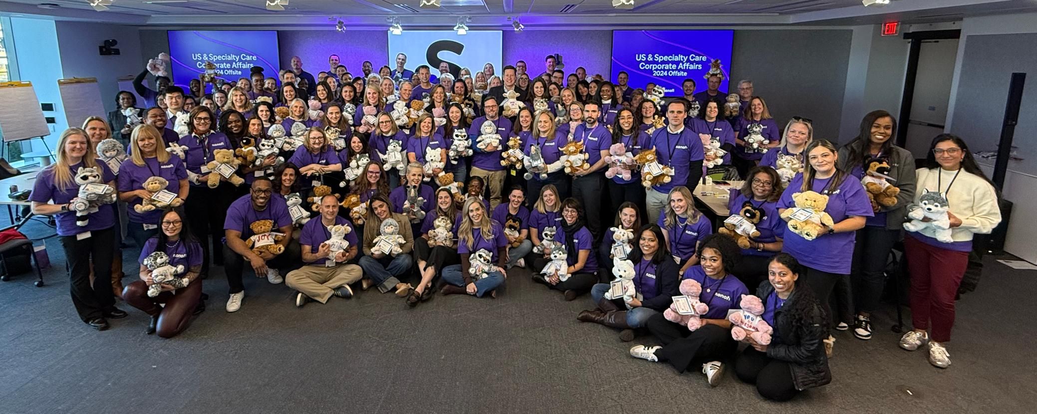 Over 100 Sanofi employees in purple t-shirts pose for a photo in a conference room while holding different kinds of stuffed animals
