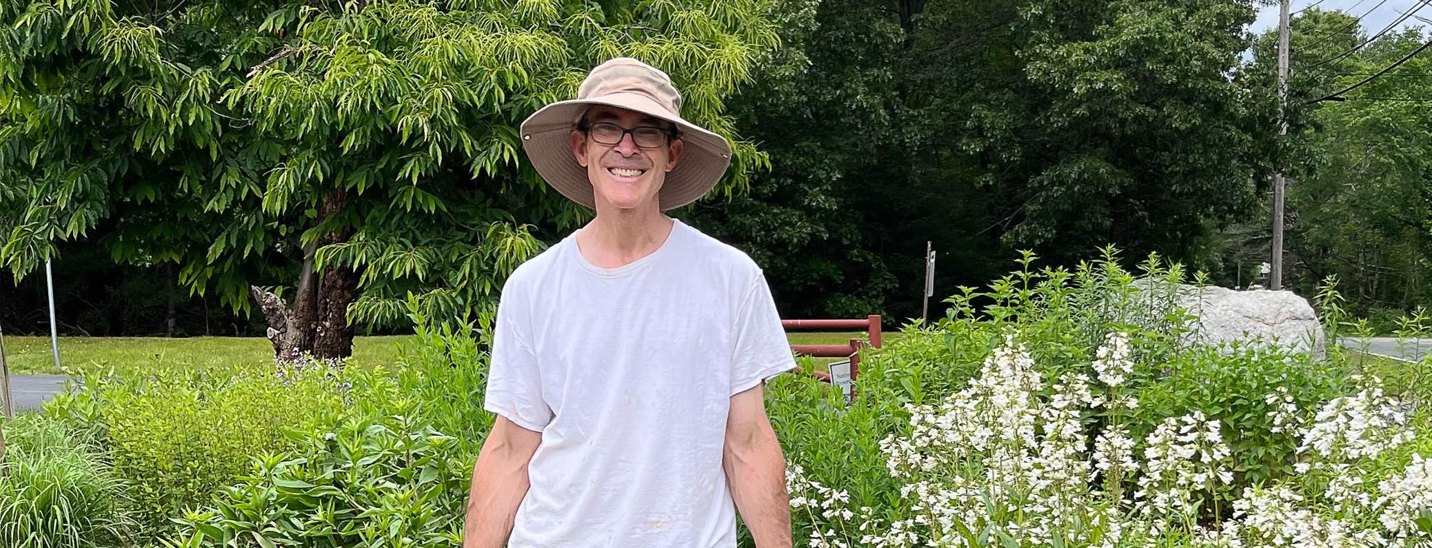Man with glasses in a gardening hat stands smiling at the camera in a community garden that he planted.