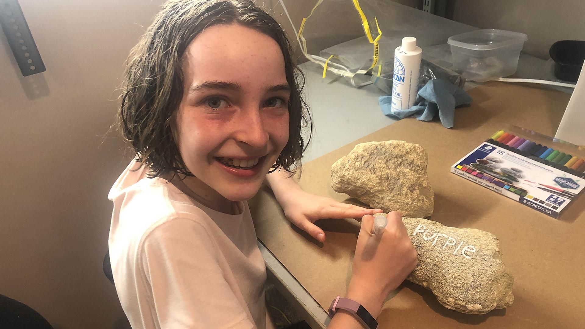 A young girl smiles at the camera as she paints the word purple onto a large rock.