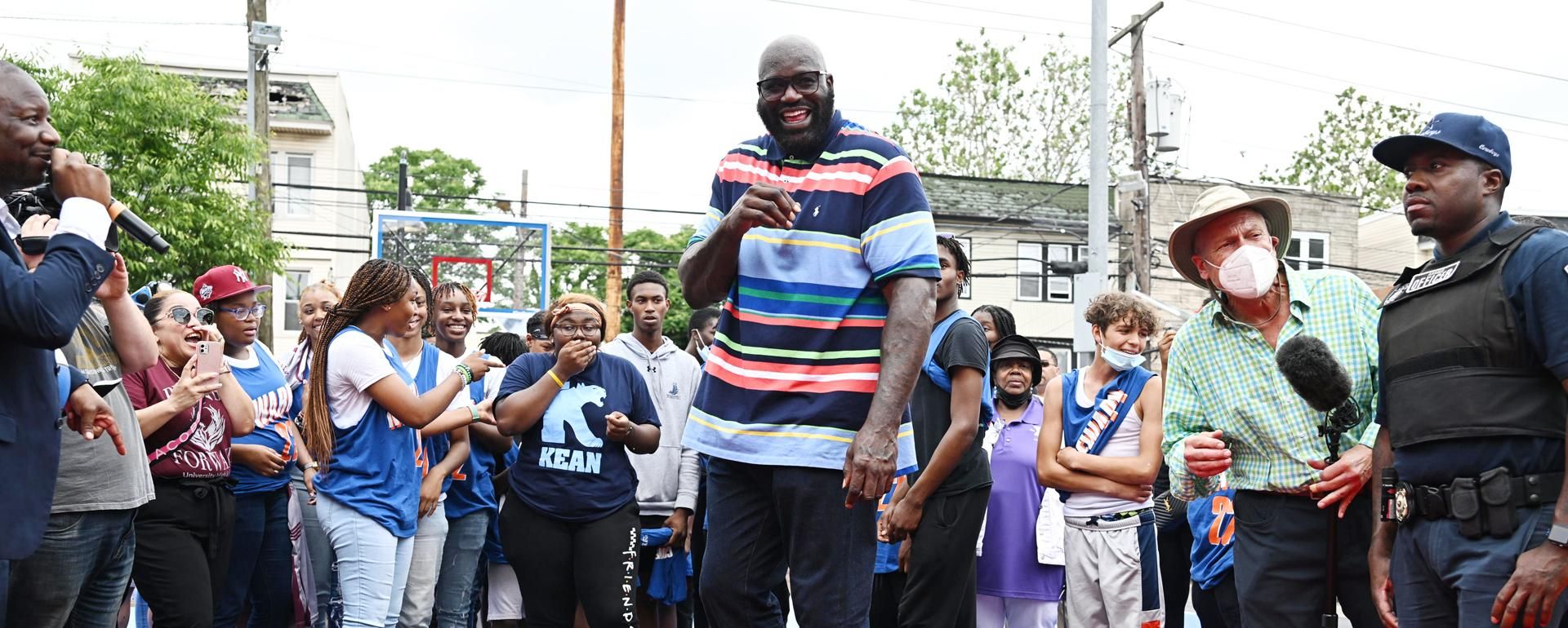 Shaquille O’Neal laughs at the camera while standing with a large groupf people of all ages at the dedication of a new basketball court.