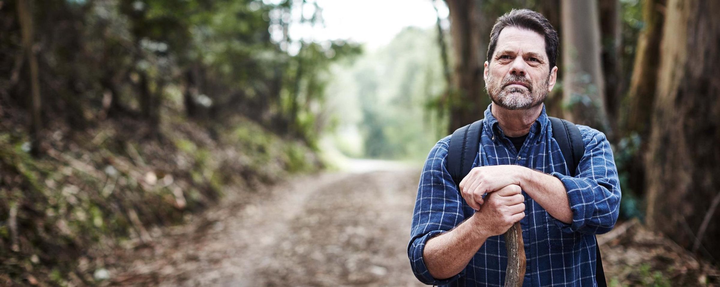 A man pausing during a walk in the woods while leaning against his walking stick.