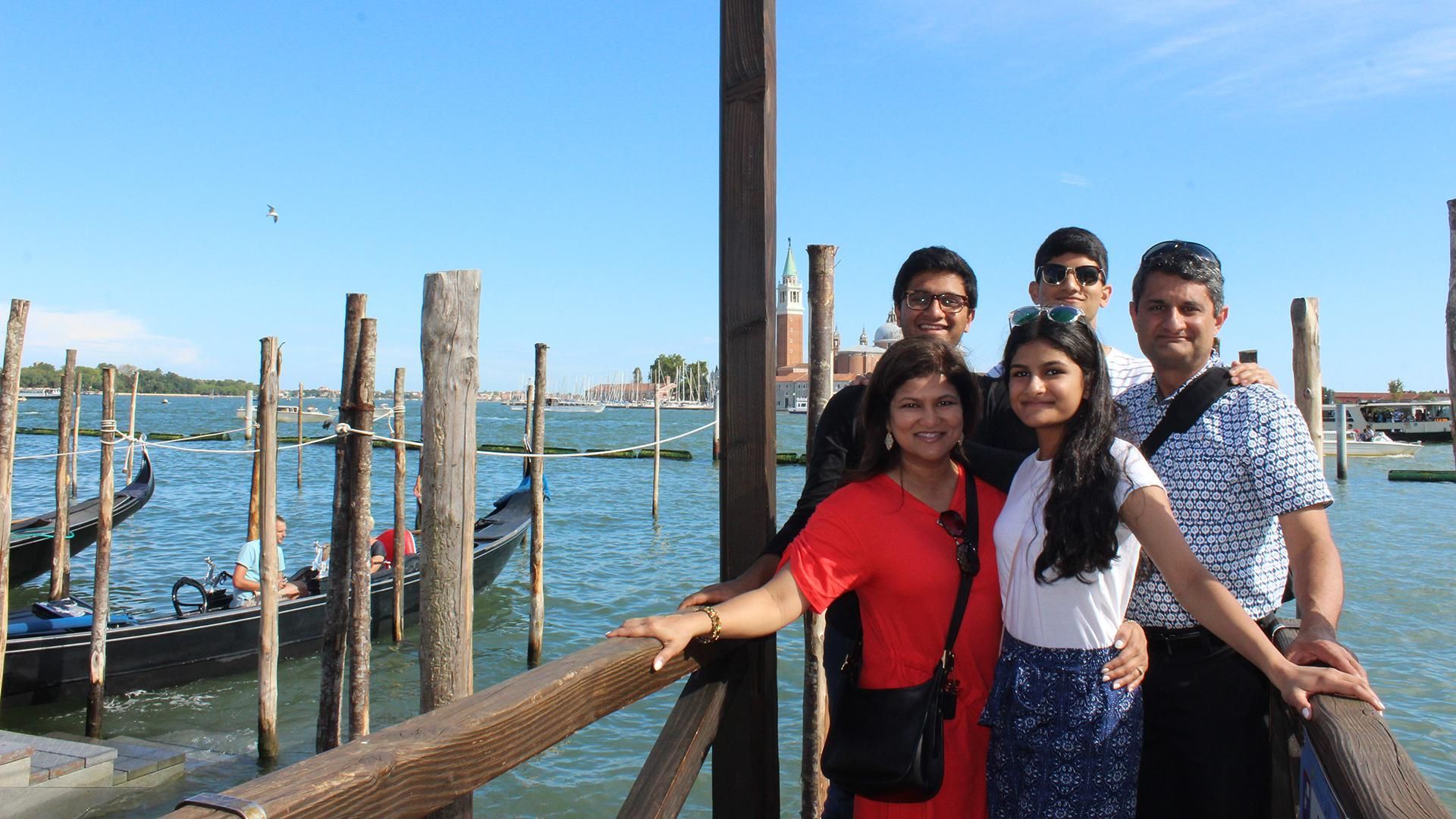 Asma Ali is standing under a lamp and on a dock in Venice, Italy with her arm around her daughter; her two sons and husband stand behind them. Gondolas are visible in the water around them.