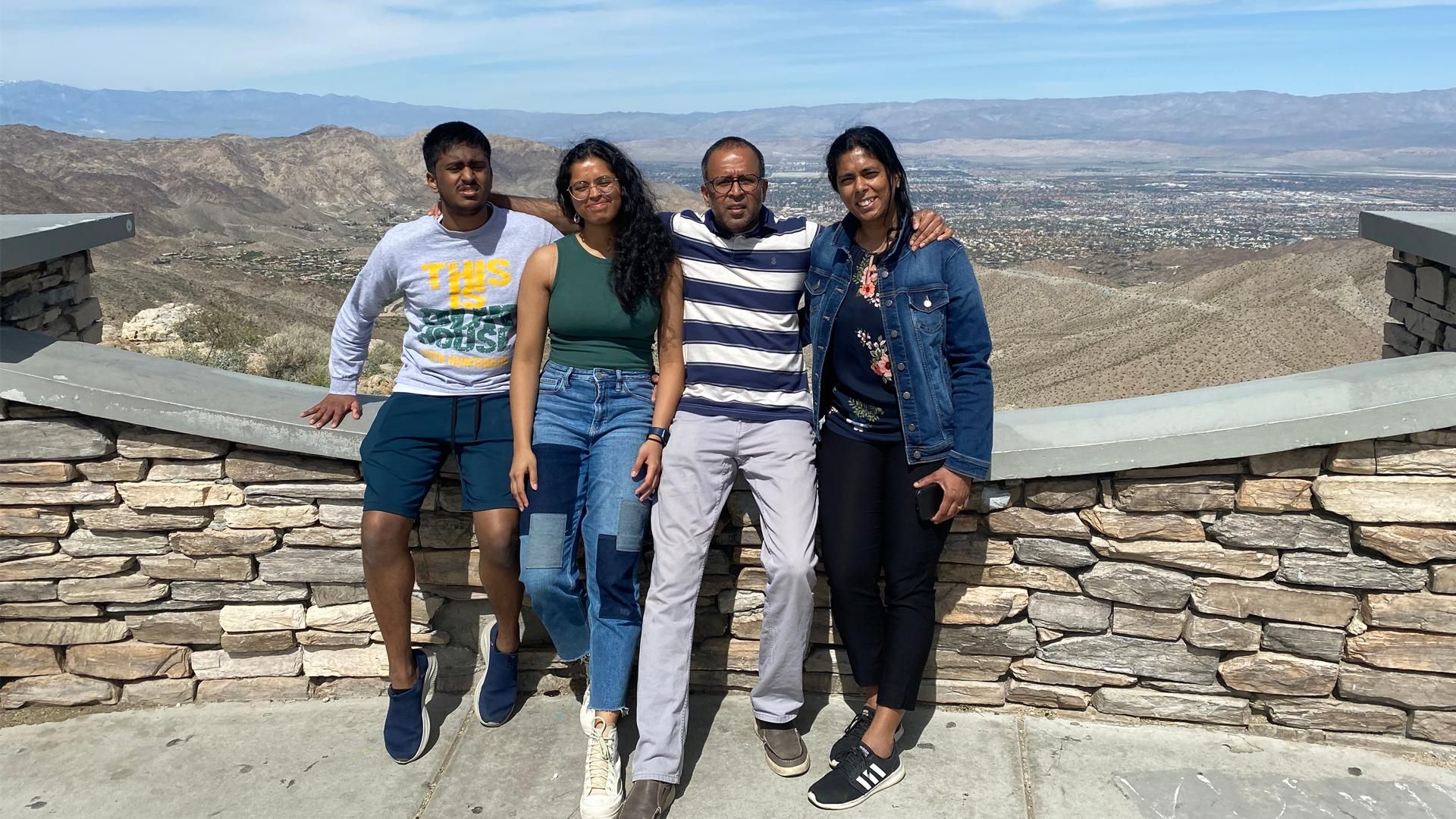 Arthi, her husband, and two children sit on a stone wall at Coachella Valley Vista Point in California on a sunny and partially cloudy afternoon.