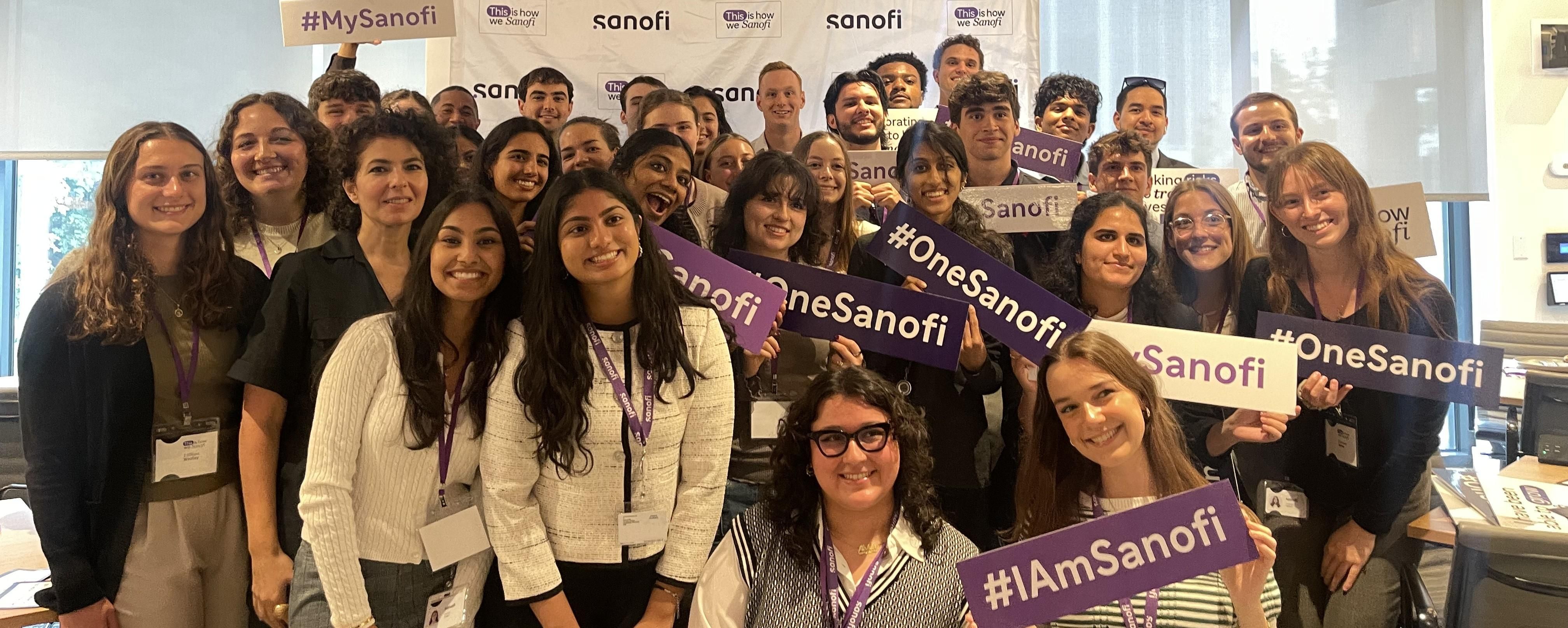 A large group of 30 to 40 interns pose, smiling at the camera in front of a Sanofi step-and-repeat. Several of them are holding signs that say “#IAmSanofi,” “#MySanofi,” or “#OneSanofi.”