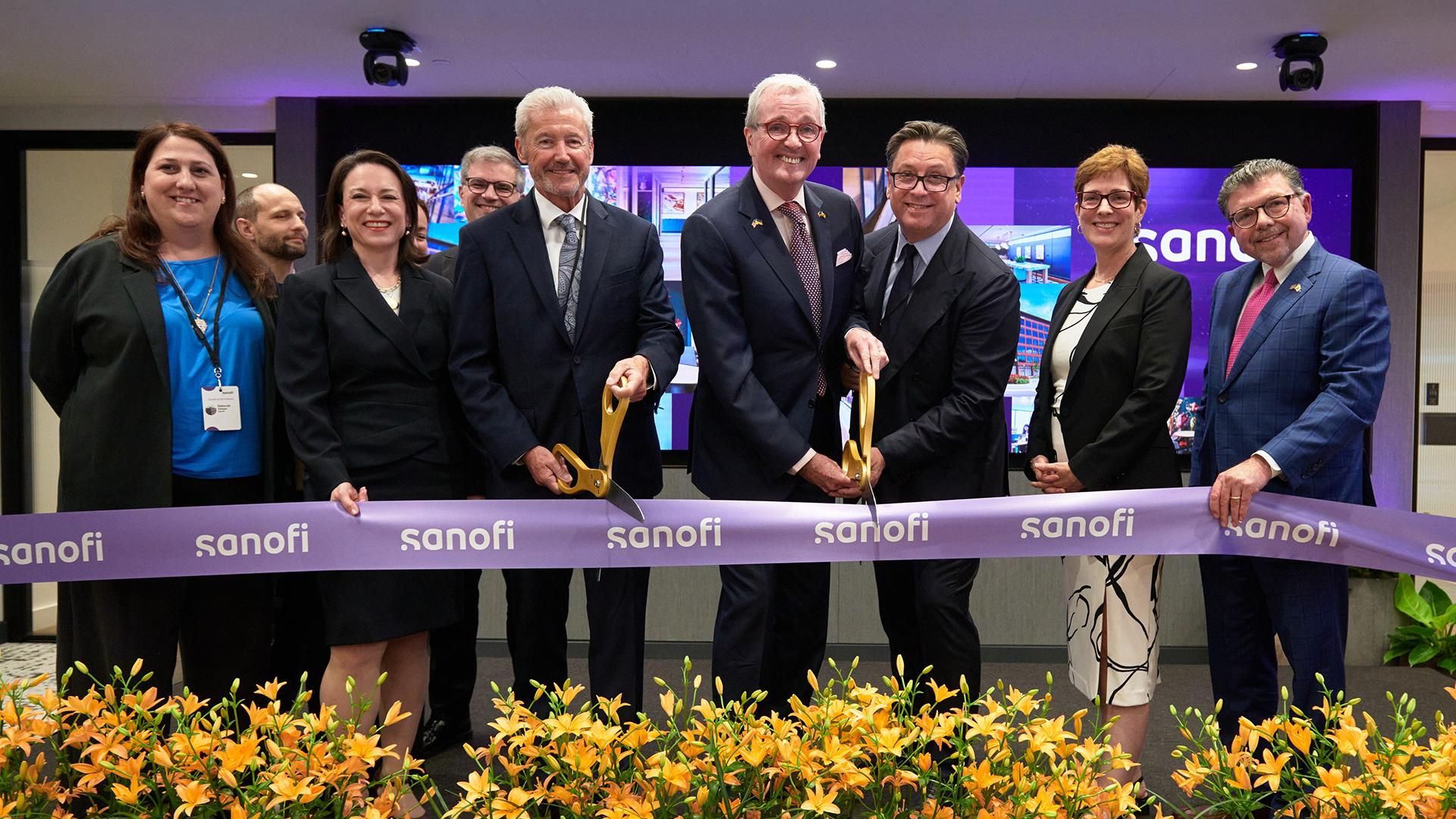 Nine people stand on a stage in a conference room cutting a purple ribbon that says Sanofi repeatedly, in front of an array of yellow flowers.