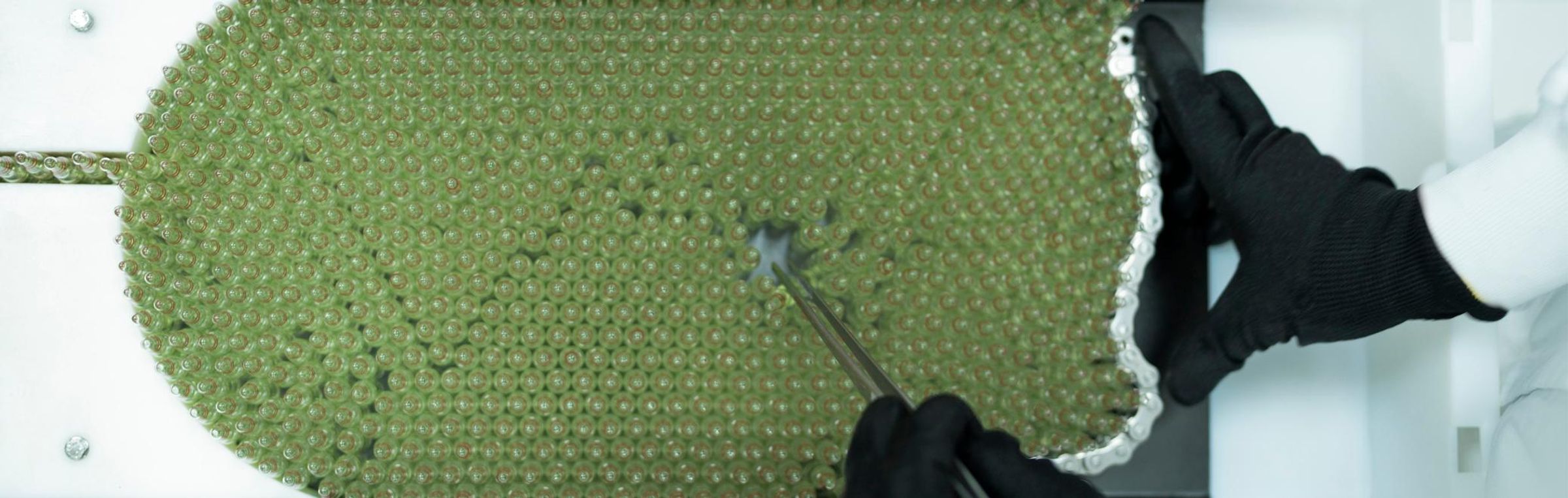 A few hundred greenish ampoules on a rack being inspected by a technician wearing black gloves and a white lab coat.