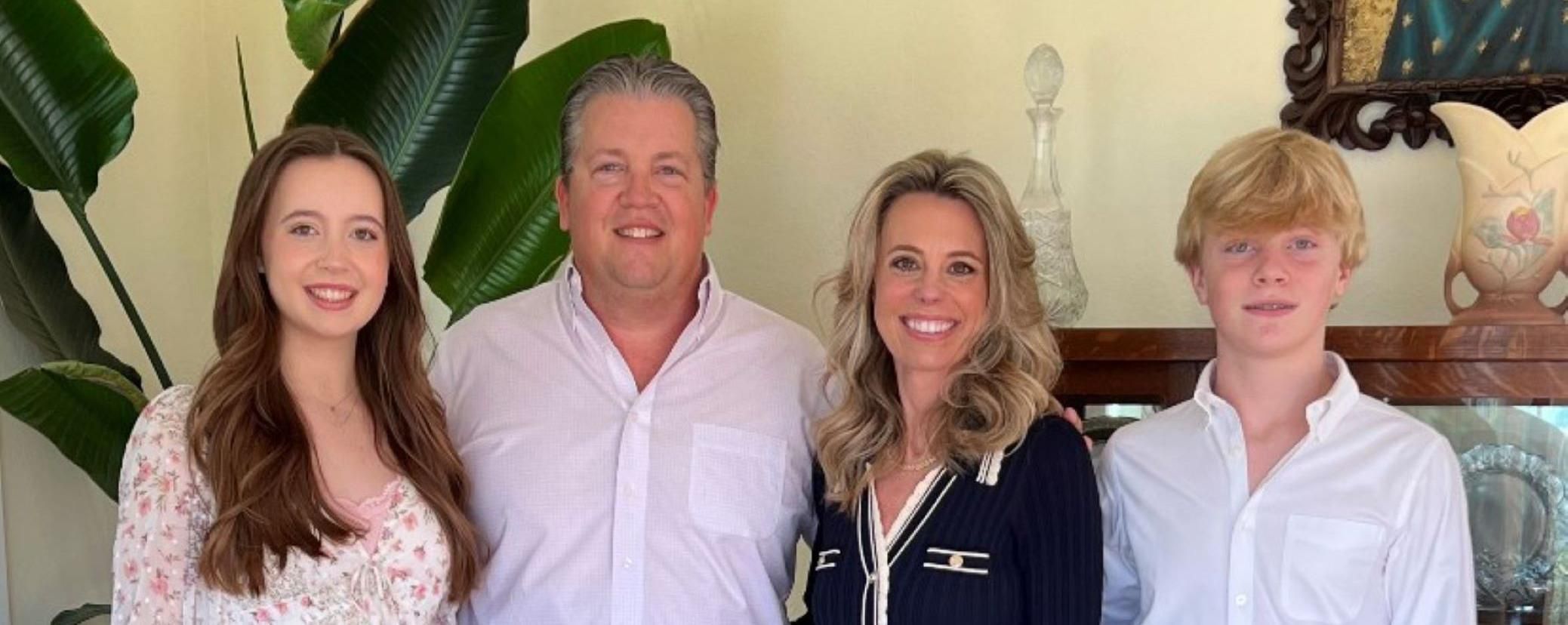 Four members of a family posing for a photo in an off-white-colored room. Family is standing in front of decorative paintings, objects, and a plant.