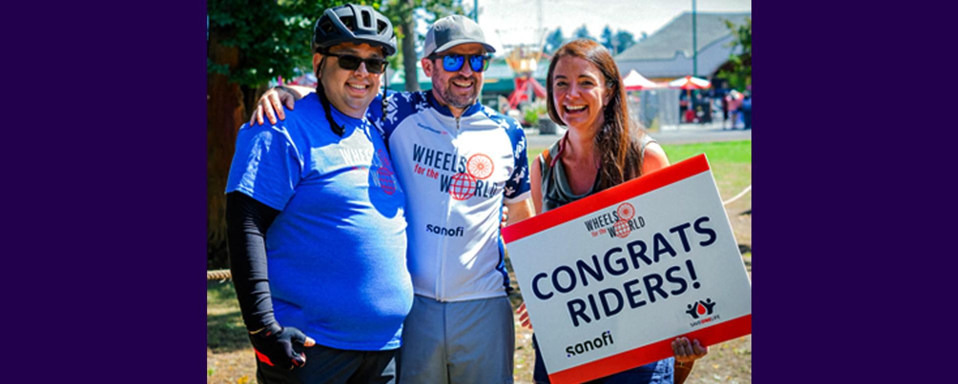 Two men in bicycle jerseys and helmets stand with a woman at the Wheels for the World event.