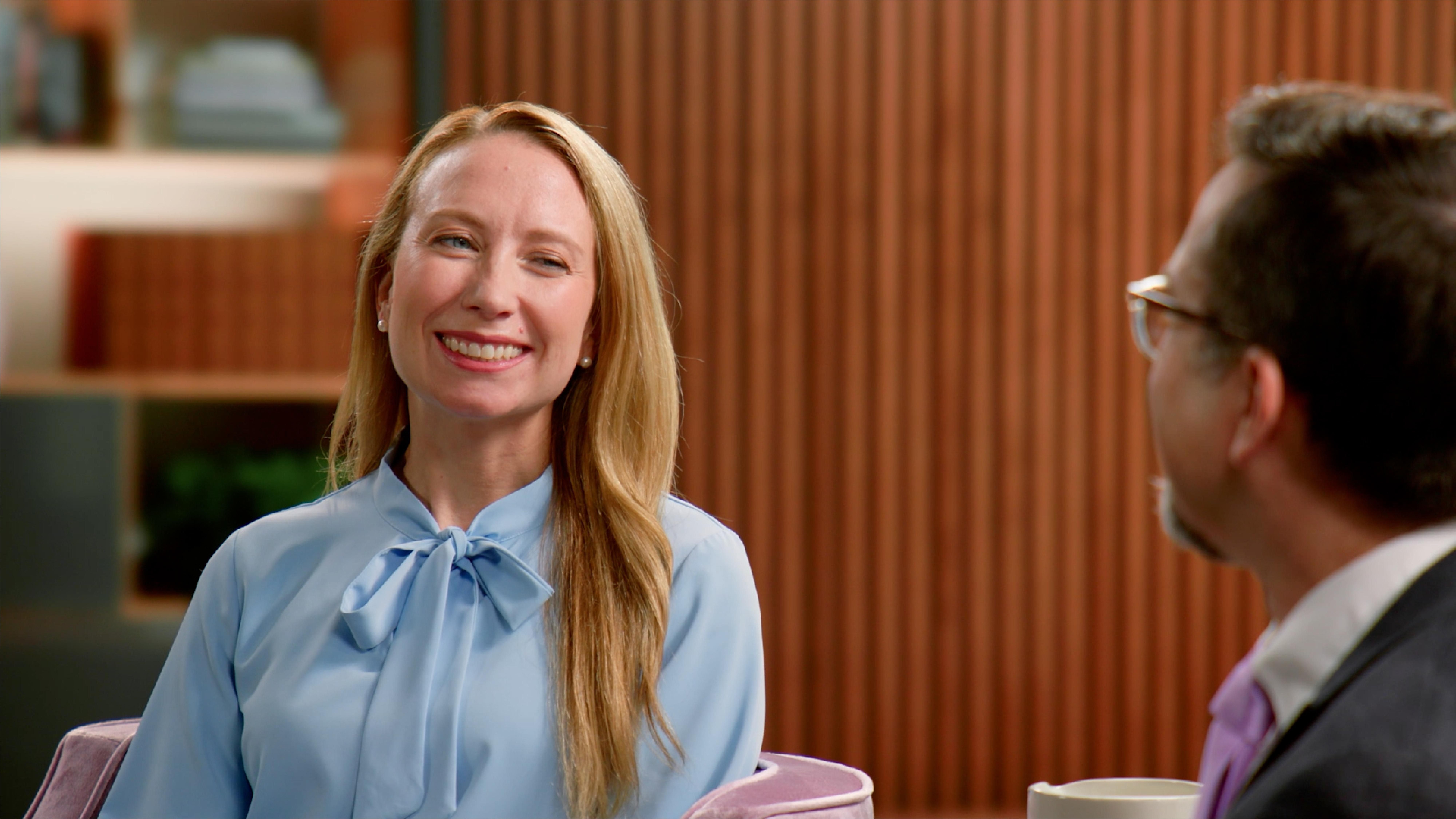 Holly, wearing a blue blouse, sits in an armchair in a living room setting, smiling at her provider, Dr. Michael Tarantino, who is wearing glasses, a grey suit, and a purple tie, to discuss her ITP management plan. 
