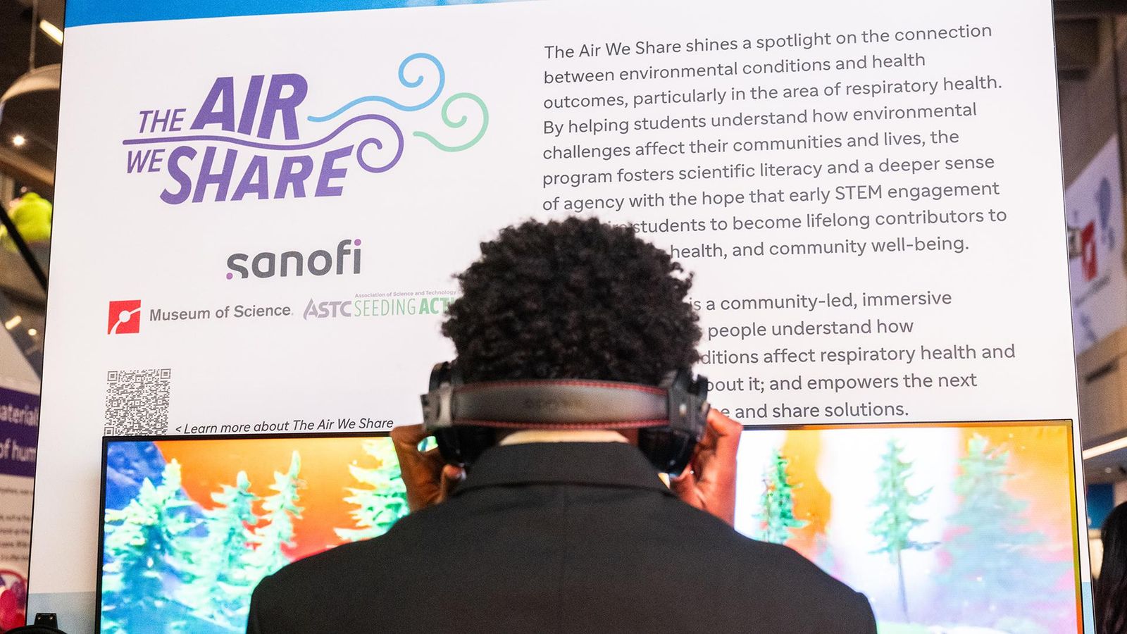 A visitor dressed in all black and wearing headphones stands before an interactive exhibit at the Museum of Science in Boston, engaging with "The Air We Share" — a community-led educational program presented by Sanofi in partnership with the Museum of Science and ASTC Seeding Action. The exhibit's central poster describes the program's mission to illuminate the connection between environmental conditions and respiratory health outcomes, foster scientific literacy among students, and empower the next generation to contribute to community well-being. A QR code in the lower left corner of the poster invites visitors to learn more. Adjacent to the poster, a monitor displays a vibrant, sunset-lit forest scene, forming part of the exhibit's immersive, interactive experience. Additional exhibits are visible in the background, situating this display within a broader event space.